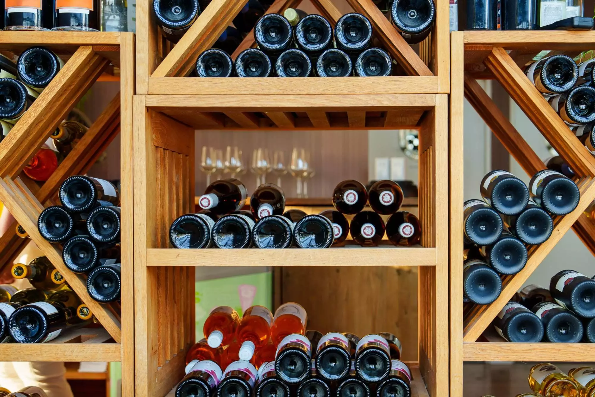 Wooden racks filled with assorted wine bottles, showcasing different colors and labels, arranged horizontally and vertically. Wine rack dimensions vary to fit the space perfectly. Three empty wine glasses are visible in the background, adding a touch of elegance to this essential wine hardware setup.