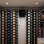 A wine cellar features wine bottles neatly stored on wall-mounted racks, with a small cooling unit centered on the back wall.