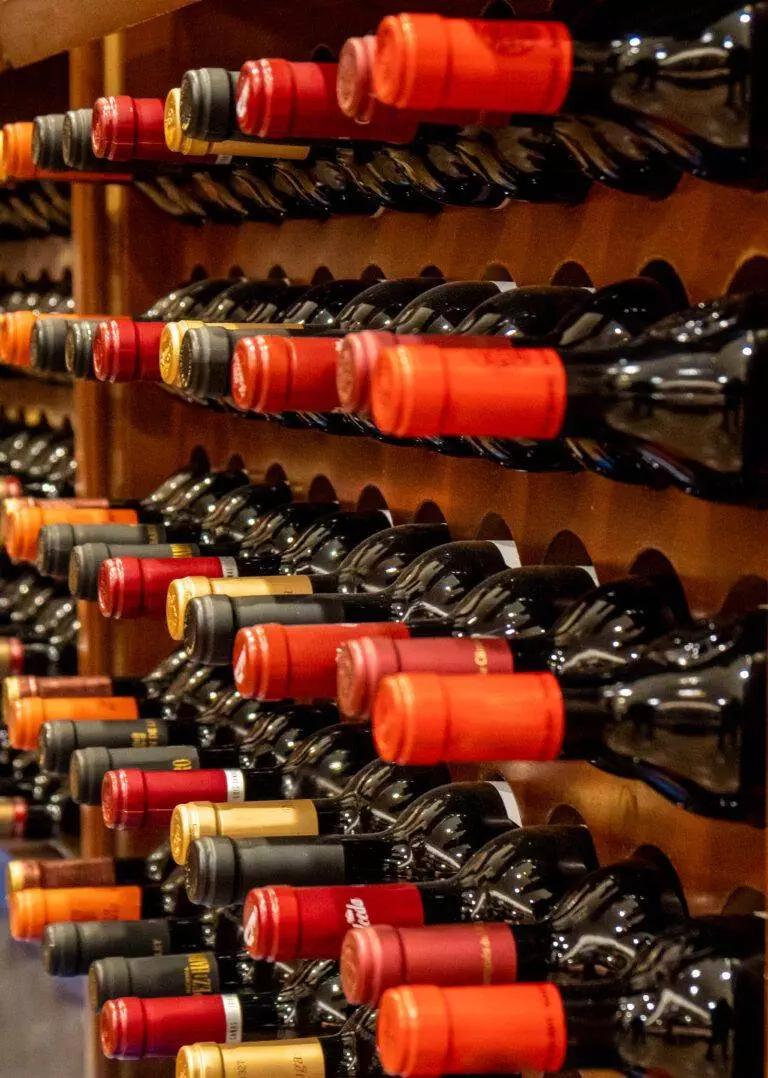 Shelves stocked with rows of wine bottles, each sealed with red or black foil caps, in a warm-lit custom wine cellar.