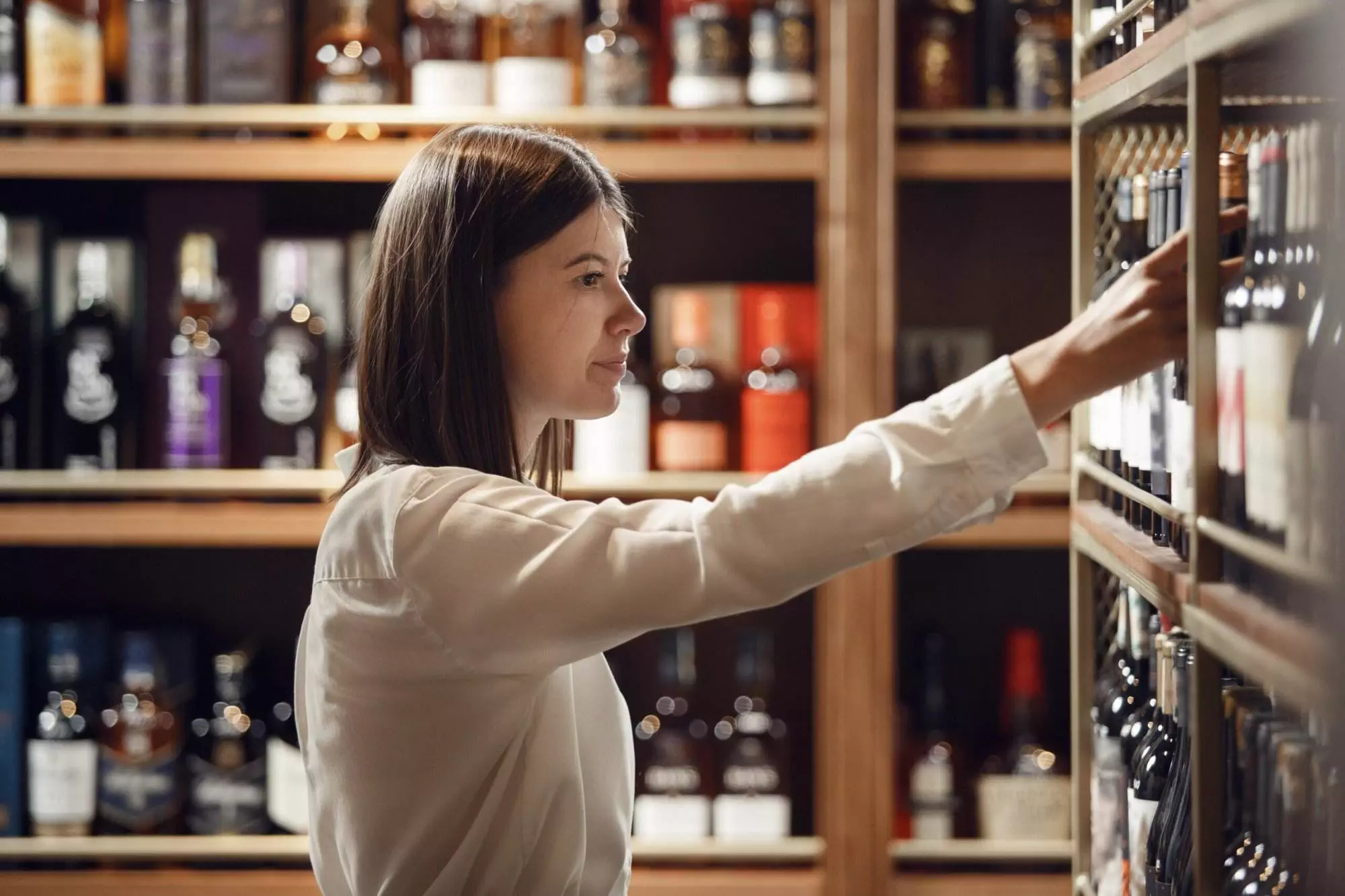 A woman wearing a white blouse reaches for a bottle on a shelf in a store, with various bottles of spirits visible in the background, all part of an impressive wine collection.