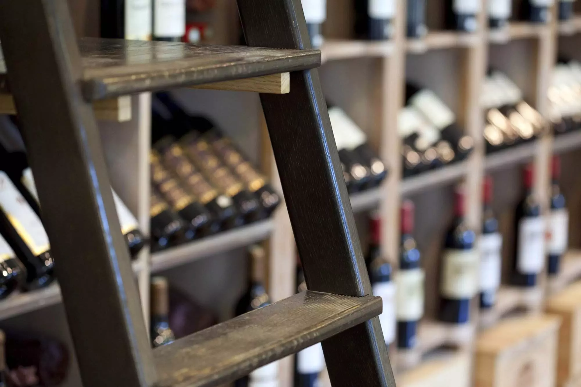 A close-up of a wooden ladder in front of a wine rack filled with various bottles of wine in a store, showcasing elegant names and premium Wine Hardware.