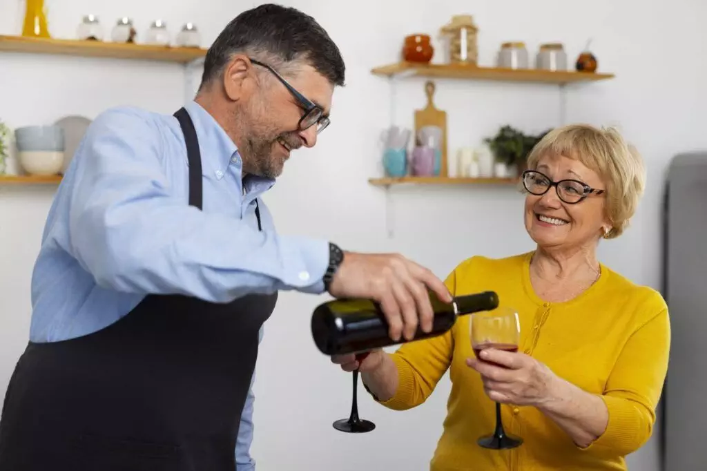 A man in an apron pours wine into a glass held by a smiling woman in a yellow shirt. They stand in a kitchen with shelves and various items, including Whisperkool wine hardware, in the background.