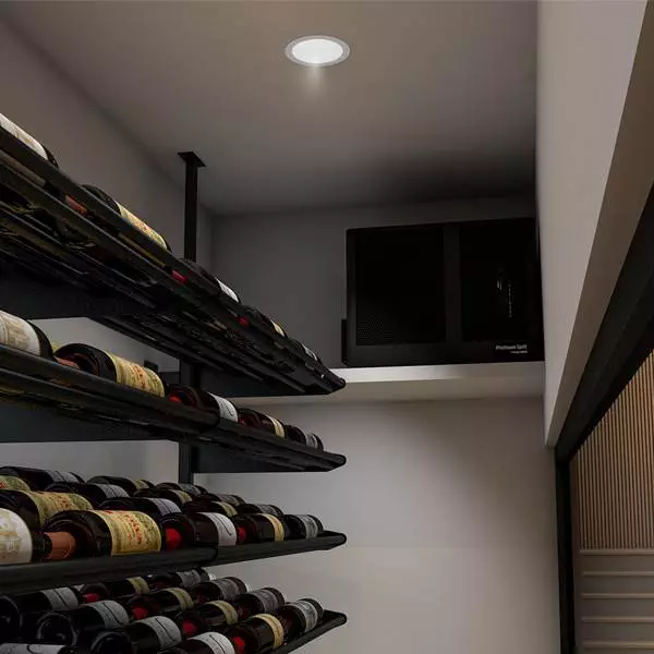 A small wine cellar with two rows of wine bottles on horizontal racks. A black electronic device is placed on a white shelf above the racks, and a round light is on the ceiling.