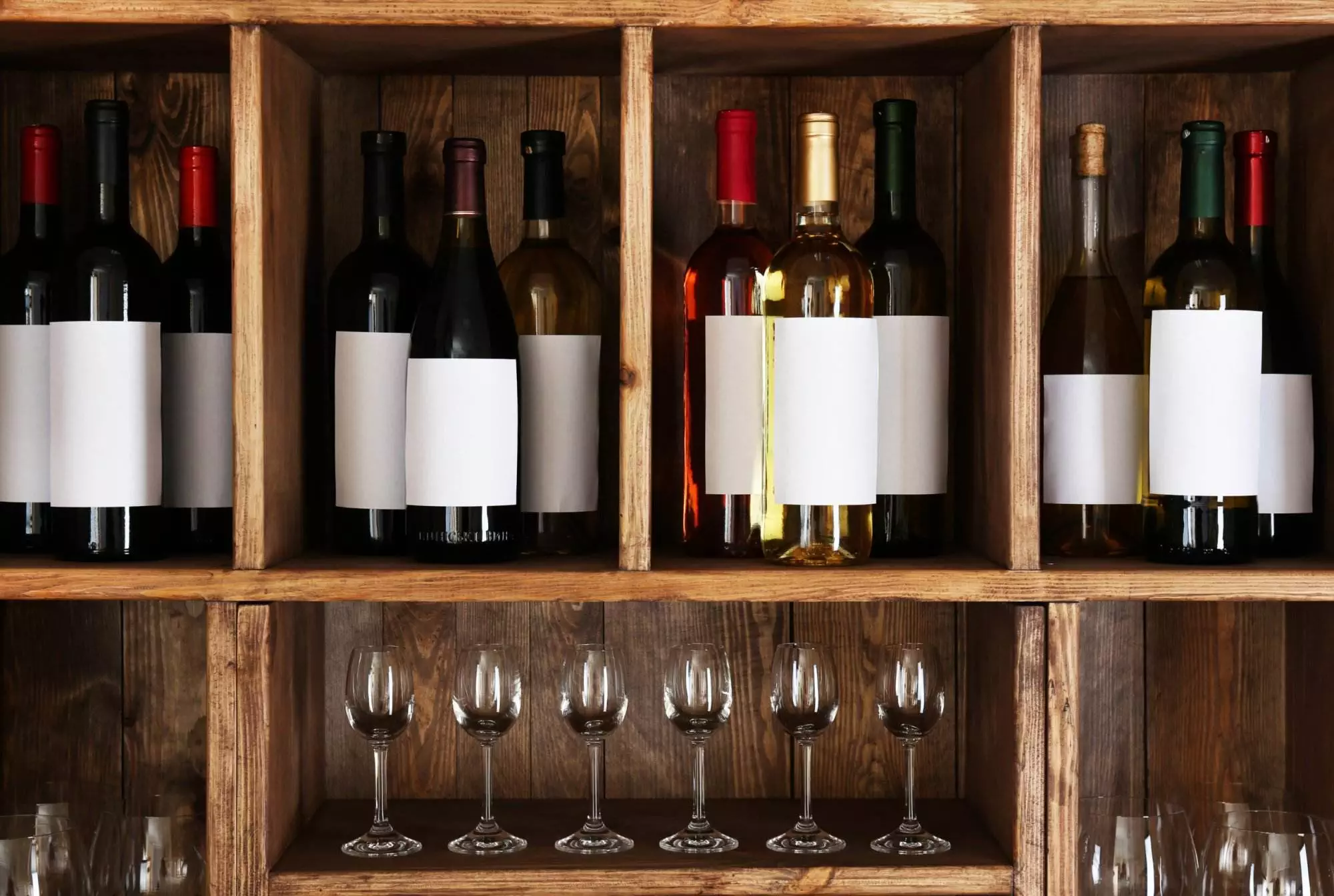 A wine rack crafted from wood showcases various wine bottles, each with blank white labels, along with several empty wine glasses neatly arranged on a lower shelf.