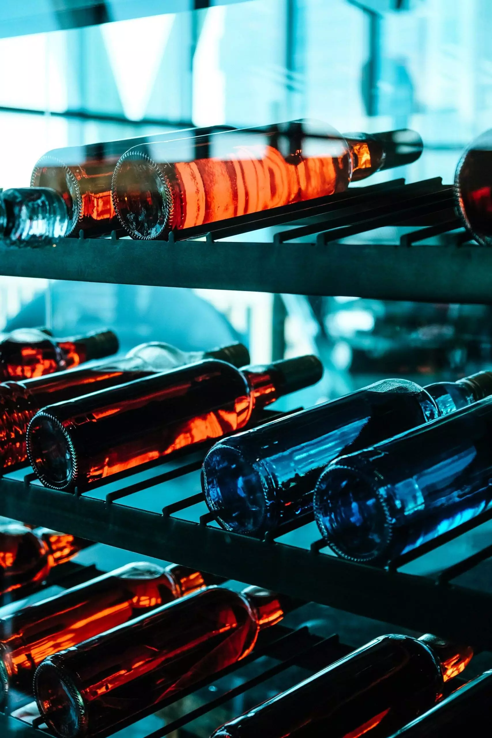 Bottles of wine, both red and blue, are stored horizontally on a sleek wine rack. The image has a cool, bluish tint due to the lighting and surroundings, offering space-saving tips through effective use of Wine Hardware.