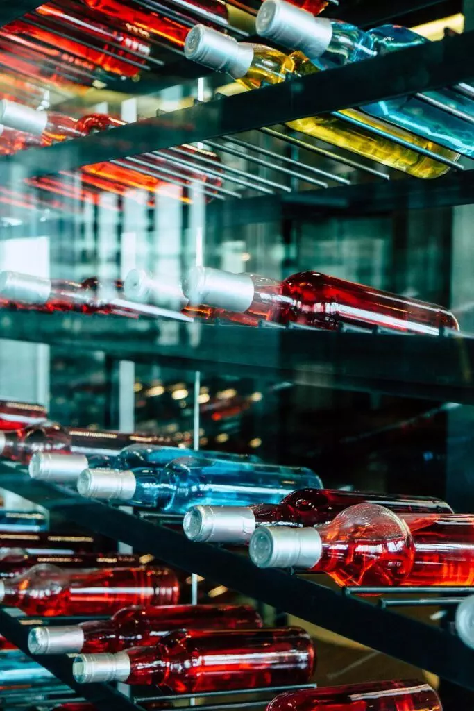 Several rows of wine bottles, filled with red, blue, and yellow liquids, are displayed on horizontal wine racks inside a glass wine cellar. This ingenious use of wine hardware ensures both elegance and efficiency while offering space-saving tips for any oenophile.