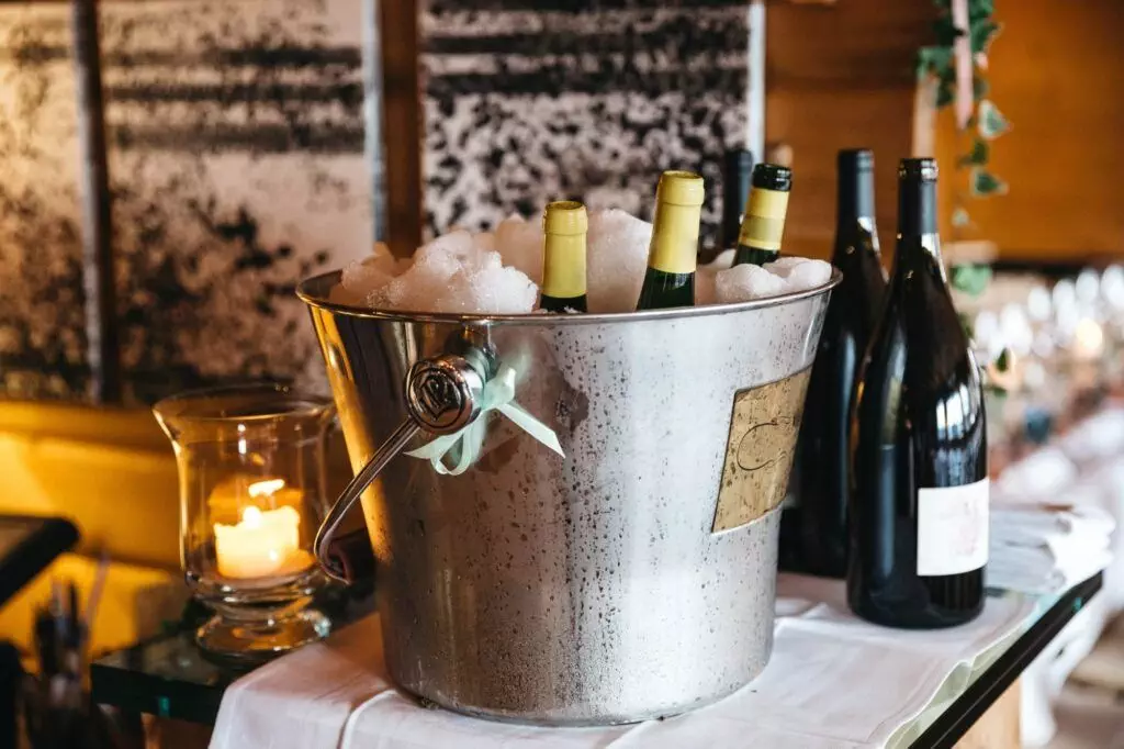 A metal bucket brimming with ice and several wine bottles sits on a table, cleverly showcasing an elegant wine storage solution for small spaces. Beside the bucket, a lit candle adds ambiance with more wine bottles nearby, completing the sophisticated display.