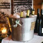 A metal bucket brimming with ice and several wine bottles sits on a table, cleverly showcasing an elegant wine storage solution for small spaces. Beside the bucket, a lit candle adds ambiance with more wine bottles nearby, completing the sophisticated display.