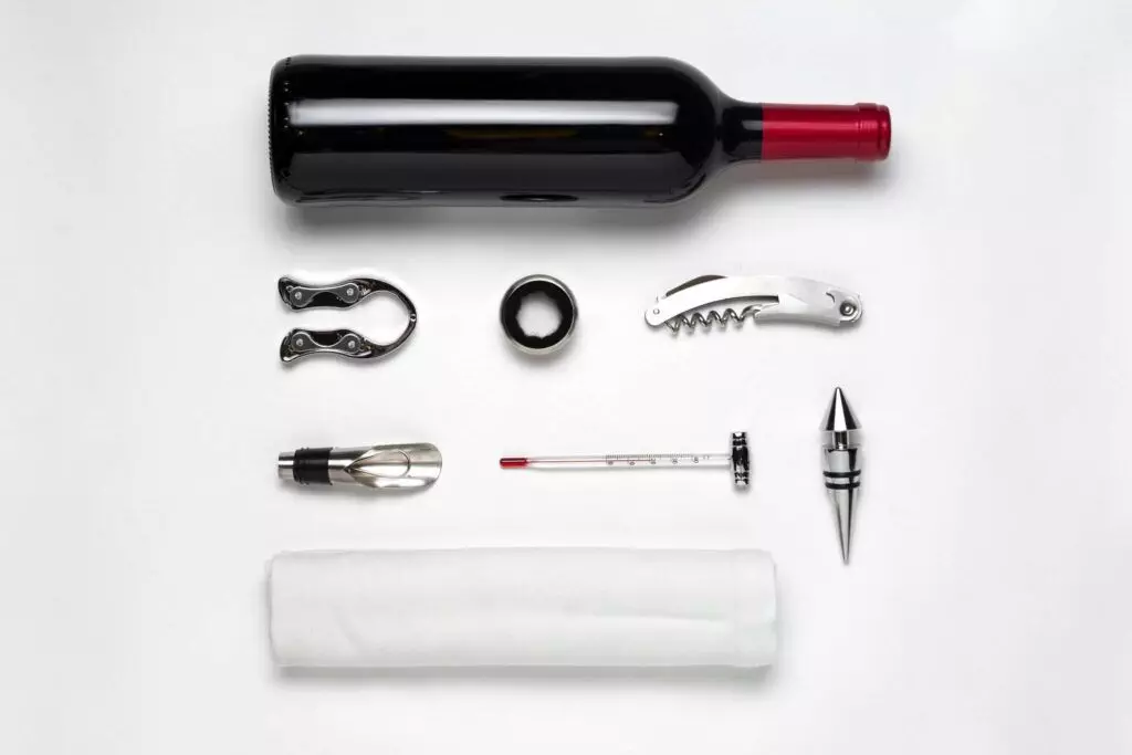 Aerial view of a bottle of red wine surrounded by must-have gadgets and tools: a corkscrew, wine stopper, foil cutter, wine aerator, wine thermometer, and a white napkin, all arranged neatly on a pristine white background.