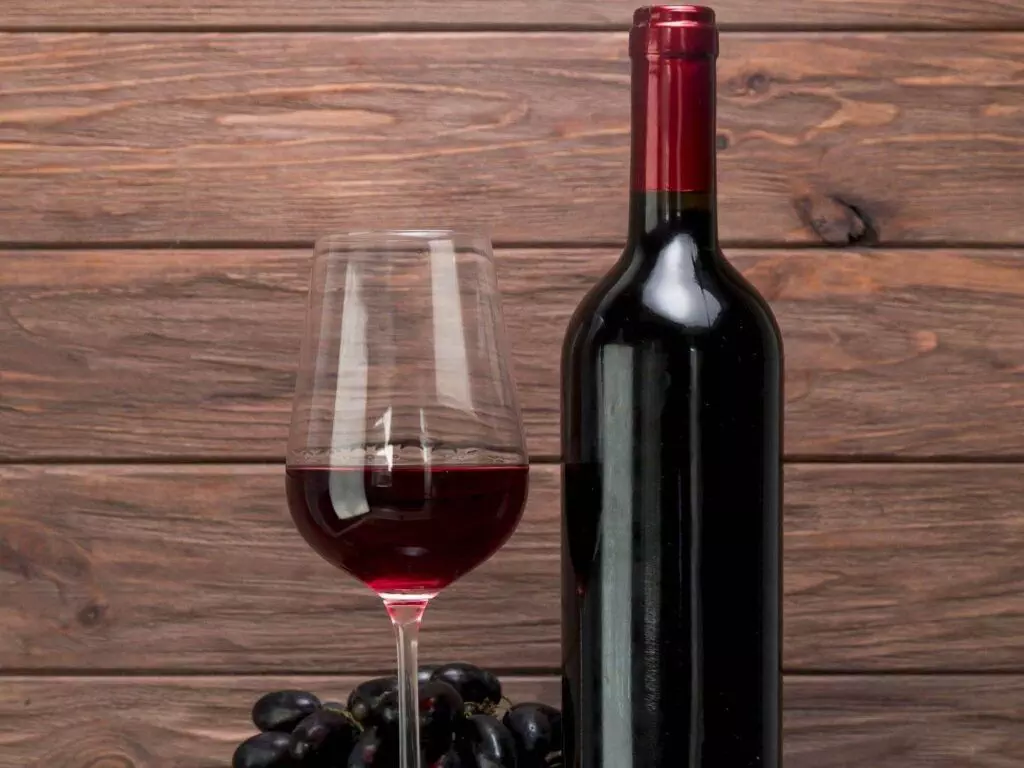 A bottle of red wine and a glass of red wine are placed on a wooden barrel with a small bunch of dark grapes and some green leaves nearby, creating an ambiance perfect for exploring your Wine Hardware collection.