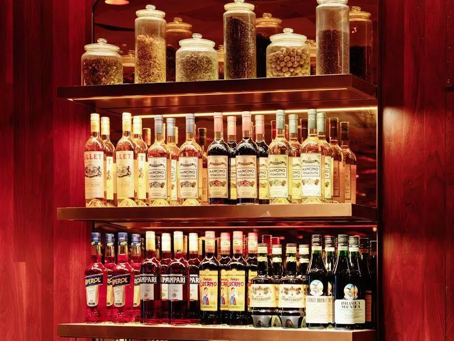 Shelves stocked with various bottles of alcohol, including wine, aperitifs, and liqueurs, in a warmly lit setting. Glass jars with dried ingredients are displayed on the top shelf.