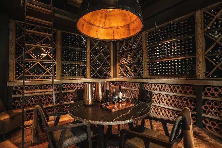 Wine cellar with wooden racks filled with bottles, a round table in the center, chairs, and a large overhead light.