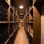 Long narrow wine cellar with walls lined with shelves full of wine bottles, dim lighting, and brick flooring.