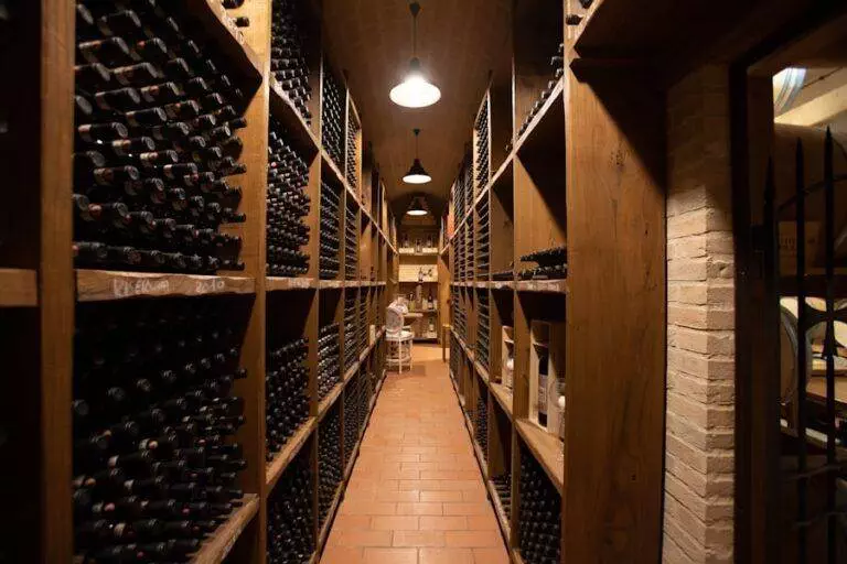 Long narrow wine cellar with walls lined with shelves full of wine bottles, dim lighting, and brick flooring.