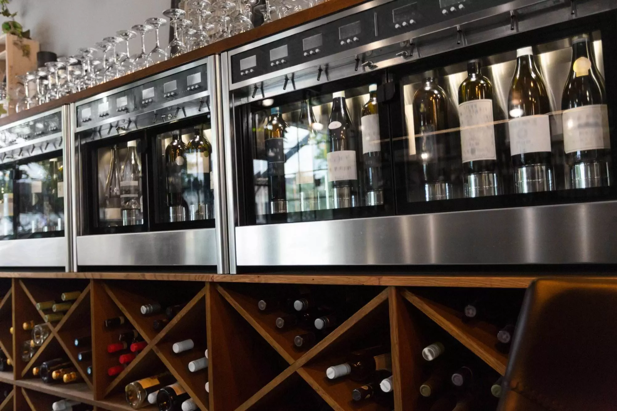 Wine bottles are showcased in a glass-fronted cooler, specifically designed for wine refrigerators, above a wooden wine rack with additional stock, creating an elegant display in this Danville bar or restaurant setting.