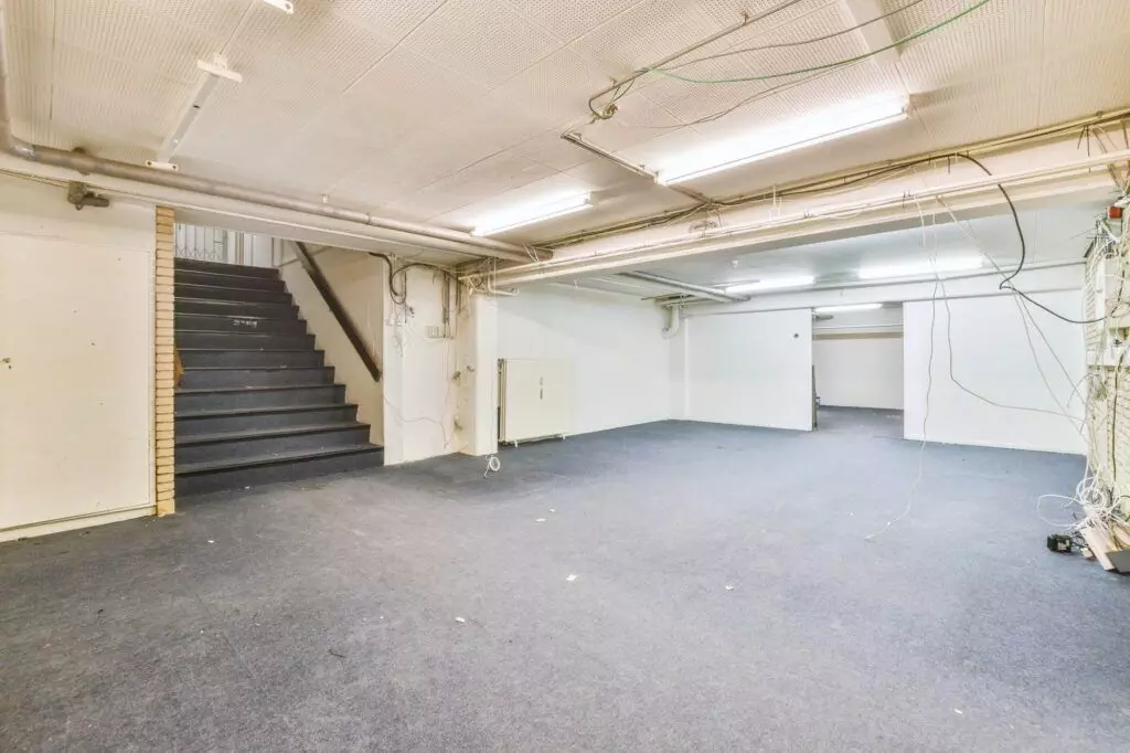 Spacious, well-lit basement conversion with carpeted floor, exposed wiring, and a staircase leading upwards. Walls are painted white, and fluorescent lights line the ceiling, perfect for repurposing unused spaces.