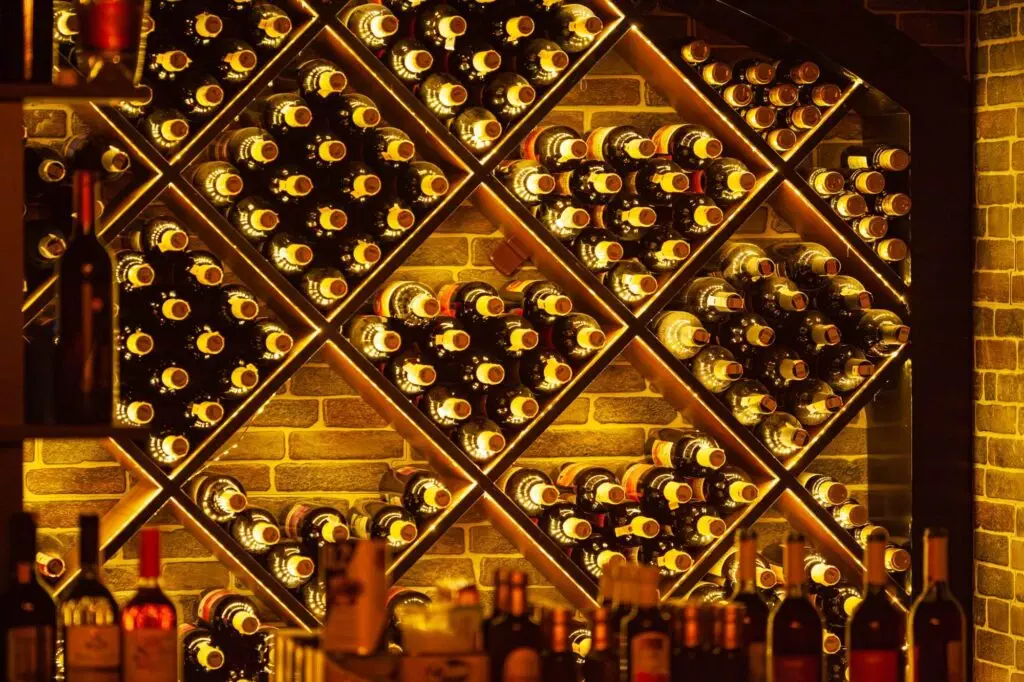 An illuminated wine cellar in Napa homes showcases a stunning array of bottles on crisscross wooden racks, enhanced by exquisite wine cellar lighting against a brick wall. Highlight your collection with this elegant and inviting display.